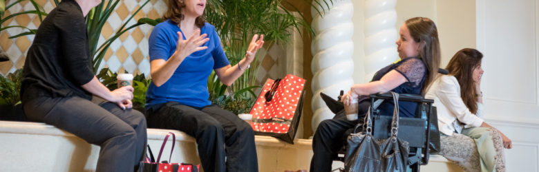 Image shows 2 women sitting next to each other, One is speaking with her hands. The other is listening. Across from them is a woman who is in a wheel chair who is also listening and holding a coffee cup. On the floor is a golden Labrador retriever service dog laying down relaxing.