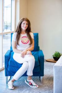 Mona Lisa Faris, publisher of DiversityComm, wearing white pants and a white Wonder Woman shirt while sitting in a blue chair. 